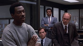 Movie still from “Beverly Hills Cop” (1984), directed by Martin Brest – A group of men in suits and ties standing in a room; Medium shot, Over the shoulder angle