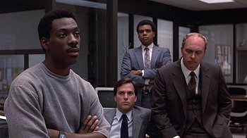 Movie still from “Beverly Hills Cop” (1984), directed by Martin Brest – A group of men in business suits sitting in a room; Medium shot, Over the shoulder angle