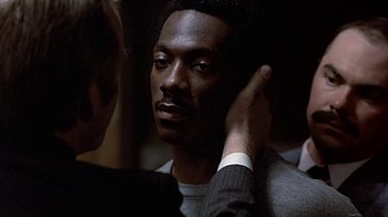 Movie still from “Beverly Hills Cop” (1984), directed by Martin Brest – A man putting his hand to his ear while another man holds his hand to his ear; Close Up shot, Over the shoulder angle