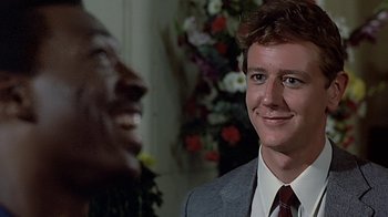 Movie still from “Beverly Hills Cop” (1984), directed by Martin Brest – A man wearing a suit and tie smiling at the camera; Close Up shot, Over the shoulder angle