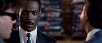 Movie still from “Beverly Hills Cop II” (1987), directed by Tony Scott – A man wearing a suit and tie standing in front of stacks of books; Close Up shot, Over the shoulder angle