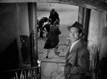 Movie still from “Bicycle Thieves” (1948), directed by Vittorio De Sica – A man in a hat standing in front of a doorway; Wide shot, High angle