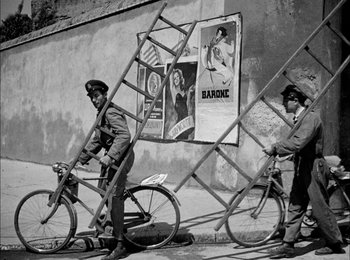 Movie still from “Bicycle Thieves” (1948), directed by Vittorio De Sica – Two men are standing next to a ladder on a bicycle; Wide shot, Low angle