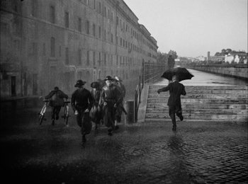 Movie still from “Bicycle Thieves” (1948), directed by Vittorio De Sica – A black and white photo of people walking in the rain with an umbrella; Wide shot, High angle