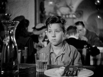 Movie still from “Bicycle Thieves” (1948), directed by Vittorio De Sica – A young boy sitting at a table with a glass of water; Medium shot, Over the shoulder angle