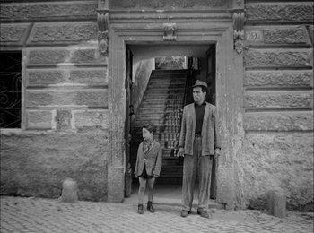 Movie still from “Bicycle Thieves” (1948), directed by Vittorio De Sica – An older man and a young boy standing in front of a building; Wide shot, Low angle