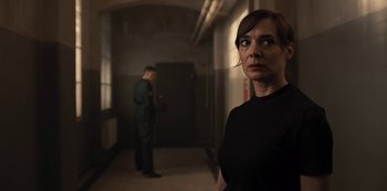 Movie still from “Dark” (2017), created by Baran bo Odar – A woman standing in a dark hallway with a man standing in the background; Medium shot, Over the shoulder angle