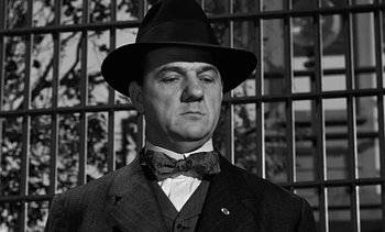 Movie still from “Birdman of Alcatraz” (1962), directed by John Frankenheimer – A man wearing a suit and a bow tie in front of a metal fence; Close Up shot, Low angle