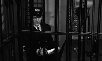 Movie still from “Birdman of Alcatraz” (1962), directed by John Frankenheimer – A man sitting in front of a jail cell holding papers; Medium shot, High angle
