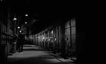 Movie still from “Birdman of Alcatraz” (1962), directed by John Frankenheimer – A man standing in a dark room next to a row of jail cell doors; Extreme Wide shot, Low angle