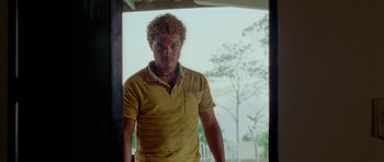 Movie still from “Birds of Passage” (2018), directed by Cristina Gallego – A man in a yellow shirt standing in front of a window; Medium shot, Low angle