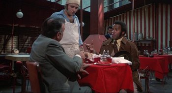 Movie still from “Black Caesar” (1973), directed by Larry Cohen – A group of men sitting at a table with food; Medium shot, Over the shoulder angle