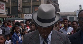 Movie still from “Black Caesar” (1973), directed by Larry Cohen – A man wearing a hat is smoking a cigarette in a crowd of people; Close Up shot, Over the shoulder angle