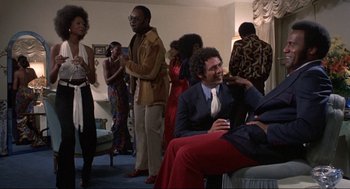 Movie still from “Black Caesar” (1973), directed by Larry Cohen – A group of people standing around a living room together; Medium shot, Low angle