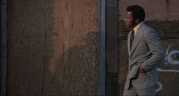 Movie still from “Black Caesar” (1973), directed by Larry Cohen – A man wearing a suit and tie standing next to a wooden wall; Medium shot, Low angle