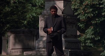 Movie still from “Black Caesar” (1973), directed by Larry Cohen – A man standing in front of a tombstone holding a book; Medium shot, Low angle