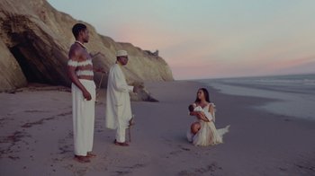 Movie still from “Black Is King” (2020), directed by Julian Klincewicz – A woman kneeling on the beach with two men; Wide shot, High angle