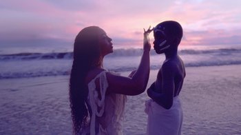Movie still from “Black Is King” (2020), directed by Julian Klincewicz – A woman and a man are painting each other's face at the beach at sunset; Medium shot, Over the shoulder angle