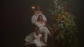 Movie still from “Black Is King” (2020), directed by Julian Klincewicz – Two women and a girl sitting in front of a flower arrangement; Wide shot, High angle
