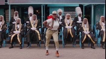 Movie still from “Black Is King” (2020), directed by Julian Klincewicz – A group of men sitting in chairs and a woman dancing; Wide shot, Low angle