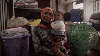 Movie still from “Black Is King” (2020), directed by Julian Klincewicz – A woman holding a baby in her arms in a room; Medium shot, Over the shoulder angle