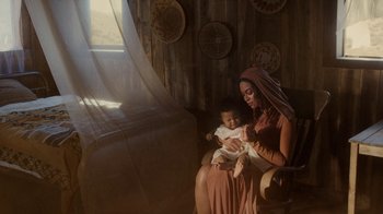 Movie still from “Black Is King” (2020), directed by Julian Klincewicz – A woman holding a baby while sitting in a chair; Medium shot, High angle