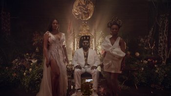 Movie still from “Black Is King” (2020), directed by Julian Klincewicz – A man sitting on top of a throne next to two women; Wide shot, Low angle