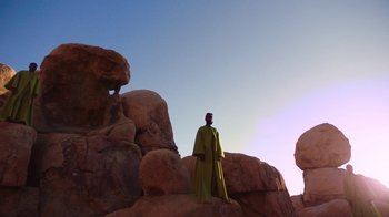 Movie still from “Black Is King” (2020), directed by Julian Klincewicz – A man standing on top of a rock formation in the desert; Extreme Wide shot, Low angle