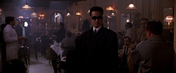Movie still from “Black Rain” (1989), directed by Ridley Scott – A man in a suit and tie standing in front of a group of diners in a restaurant; Medium shot, Over the shoulder angle