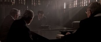 Movie still from “Black Rain” (1989), directed by Ridley Scott – A man in a suit and tie sitting at a table in front of a cell phone; Medium shot, Low angle