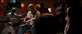 Movie still from “Black Snake Moan” (2006), directed by Craig Brewer – A man playing a guitar in front of a group of people; Medium shot, Over the shoulder angle