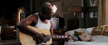 Movie still from “Black Snake Moan” (2006), directed by Craig Brewer – An older man playing a guitar in a living room; Close Up shot, Over the shoulder angle