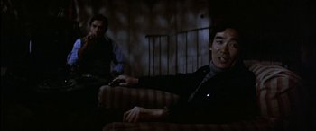 Movie still from “Black Sunday” (1977), directed by John Frankenheimer – A man sitting on top of a couch next to another man holding a remote; Medium shot, Over the shoulder angle