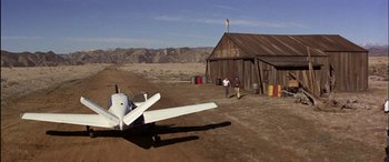 Movie still from “Black Sunday” (1977), directed by John Frankenheimer – An airplane sitting on top of a dirt field near a building; Extreme Wide shot, Low angle