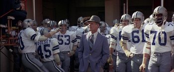 Movie still from “Black Sunday” (1977), directed by John Frankenheimer – A man in a suit and tie standing in front of a group of football players; Medium shot, Low angle