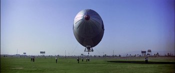 Movie still from “Black Sunday” (1977), directed by John Frankenheimer – A group of people standing in the grass next to an airship; Extreme Wide shot, Low angle