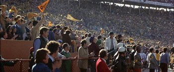 Movie still from “Black Sunday” (1977), directed by John Frankenheimer – A group of people standing in a crowd at a stadium; Extreme Wide shot, High angle