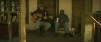 Movie still from “Blaze” (2018), directed by Ethan Hawke – A man playing a guitar while sitting next to another man holding a cup of coffee; Medium shot, Over the shoulder angle