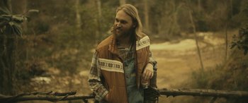 Movie still from “Blaze” (2018), directed by Ethan Hawke – A man with long hair and a beard is leaning against a fence; Medium shot, Over the shoulder angle