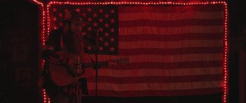 Movie still from “Blaze” (2018), directed by Ethan Hawke – A person holding a guitar in front of an american flag; Wide shot, Low angle