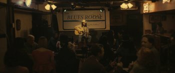 Movie still from “Blaze” (2018), directed by Ethan Hawke – A man in a cowboy hat playing a guitar in front of an audience; Wide shot, Over the shoulder angle