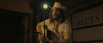 Movie still from “Blaze” (2018), directed by Ethan Hawke – A man in a cowboy hat playing a guitar; Medium shot, Over the shoulder angle