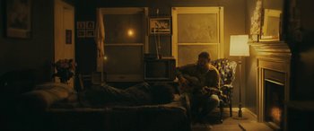 Movie still from “Blaze” (2018), directed by Ethan Hawke – A man sitting on a couch playing a guitar; Wide shot, Over the shoulder angle