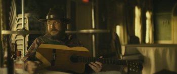 Movie still from “Blaze” (2018), directed by Ethan Hawke – A man with a beard and a hat is playing a guitar; Close Up shot, Over the shoulder angle