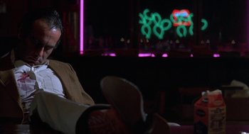 Movie still from “Blood Simple” (1984), directed by Joel Coen – A man sitting on a couch in front of a neon sign; Medium shot, Low angle