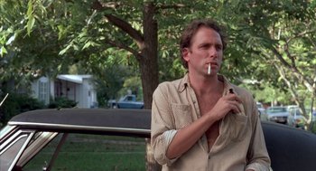 Movie still from “Blood Simple” (1984), directed by Joel Coen – A man smoking a cigarette while standing next to a tree; Medium shot, Low angle
