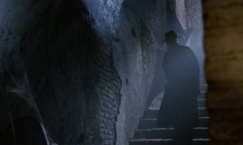 Movie still from “Blood and Black Lace” (1964), directed by Mario Bava – A man wearing a black hat and a black coat walking up some stairs; Wide shot, Low angle
