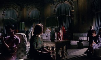 Movie still from “Blood and Black Lace” (1964), directed by Mario Bava – A woman sitting on a chair in front of a table; Wide shot, Over the shoulder angle