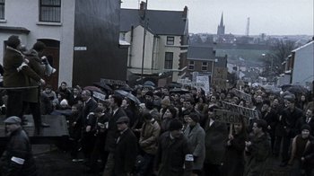 Movie still from “Bloody Sunday” (2002), directed by Paul Greengrass – A large group of people are holding signs and umbrellas; Extreme Wide shot, High angle