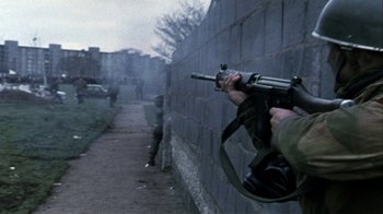 Movie still from “Bloody Sunday” (2002), directed by Paul Greengrass – A man holding a gun while standing next to a brick wall; Wide shot, Over the shoulder angle